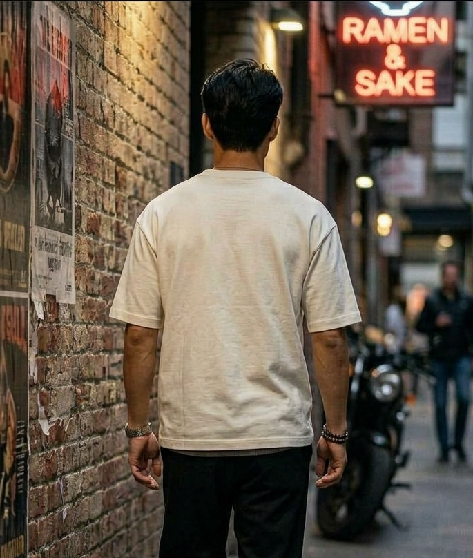 A man viewed from behind wearing a light beige crewneck t-shirt and black pants, standing in a narrow city alleyway with a brick wall on the left and a neon sign reading 'RAMEN & SAKE' on the right.